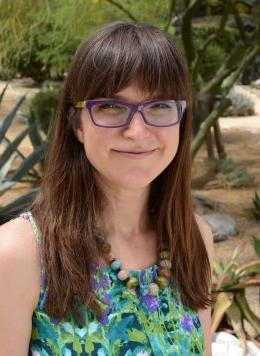 Woman with brown hair, wearing glasses, and wearing a green shirt and beaded necklace standing outdoors.